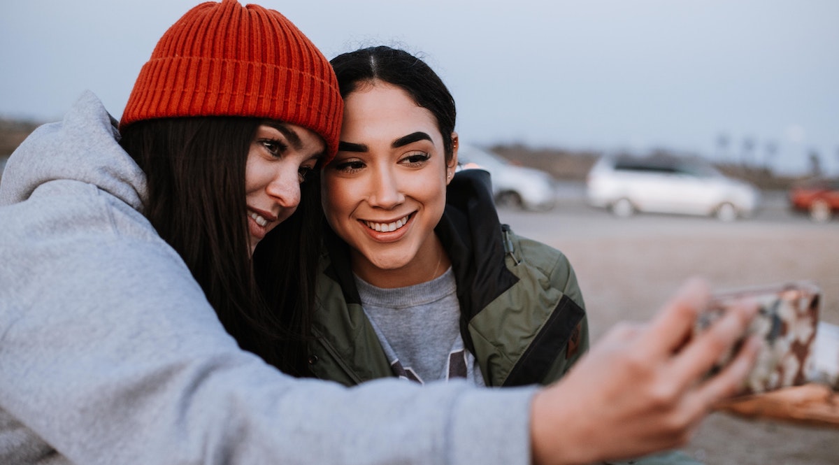 two female presenting young people heads together posing for selfie outside in parking lot